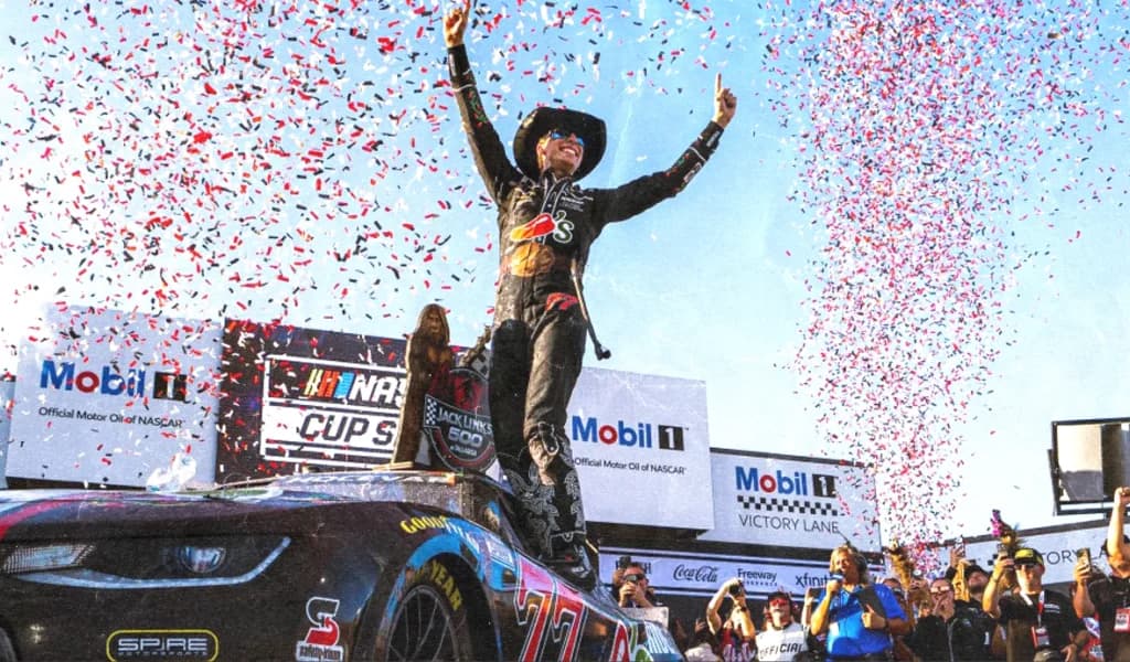 A NASCAR driver celebrates on top of his car in victory lane as confetti flies.