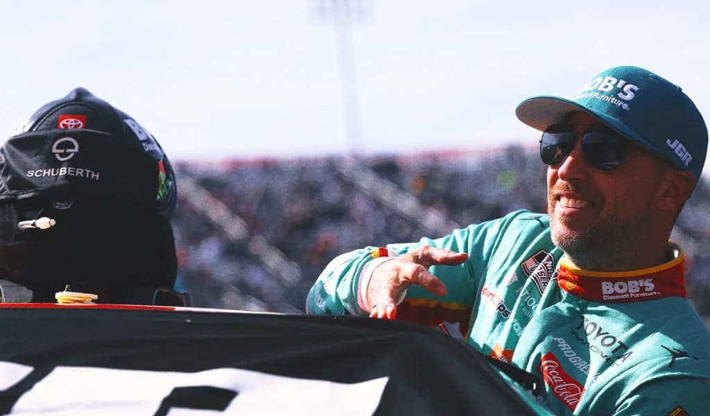 A NASCAR driver in a teal firesuit and sunglasses smiles at the track.