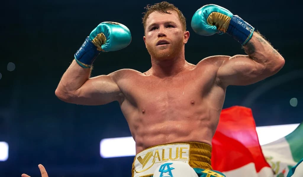 Canelo Alvarez raises his gloves in the ring with the Mexican flag behind him.