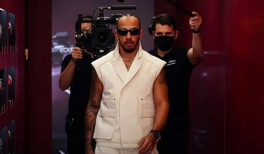 Lewis Hamilton in an all-white outfit walks the paddock trailed by a camera crew.